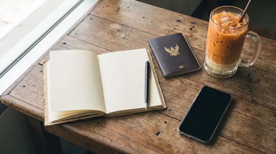 Wooden desk with a blank notebook, a pen, a smartphone, a passport and a cup of Thai iced tea in soft light