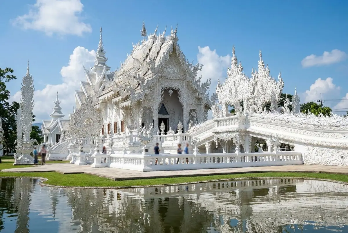 The intricate white and mirrored architecture of Wat Rong Khun, the White Temple, over a pond under blue sky
