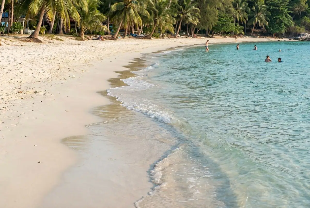 Calm white-sand beach on Koh Samet with turquoise shallow water, palm trees and a few swimmers