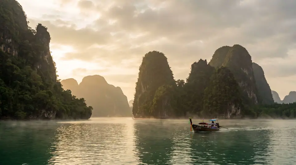 Limestone karst islands rising from calm emerald water in Phang Nga Bay at sunrise, a long-tail boat below